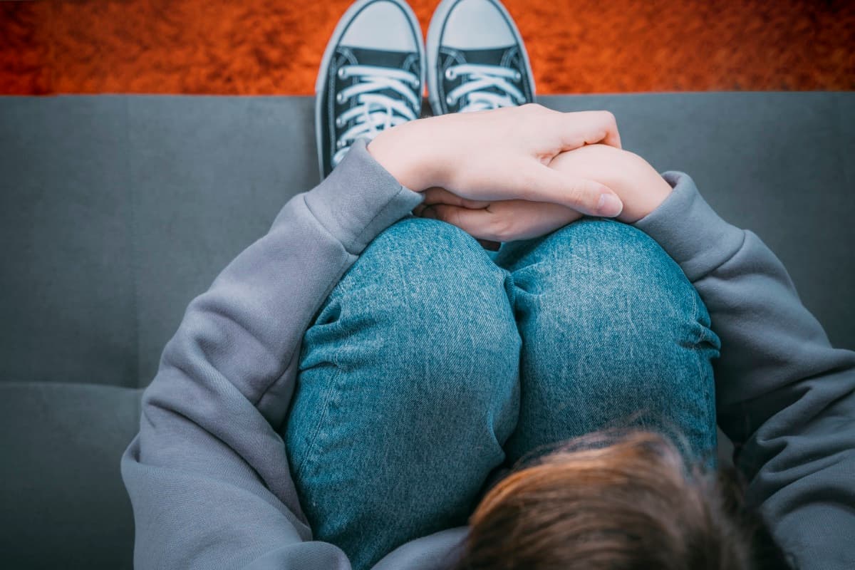 Junger Mensch im grauen Pullover sitzt zusammengekauert auf einem Sofa und umschließt die Knie mit den Händen